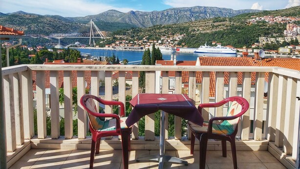 Un balcón con dos sillas y una mesa, con vistas a un pueblo costero con montañas, un puente y un barco.
