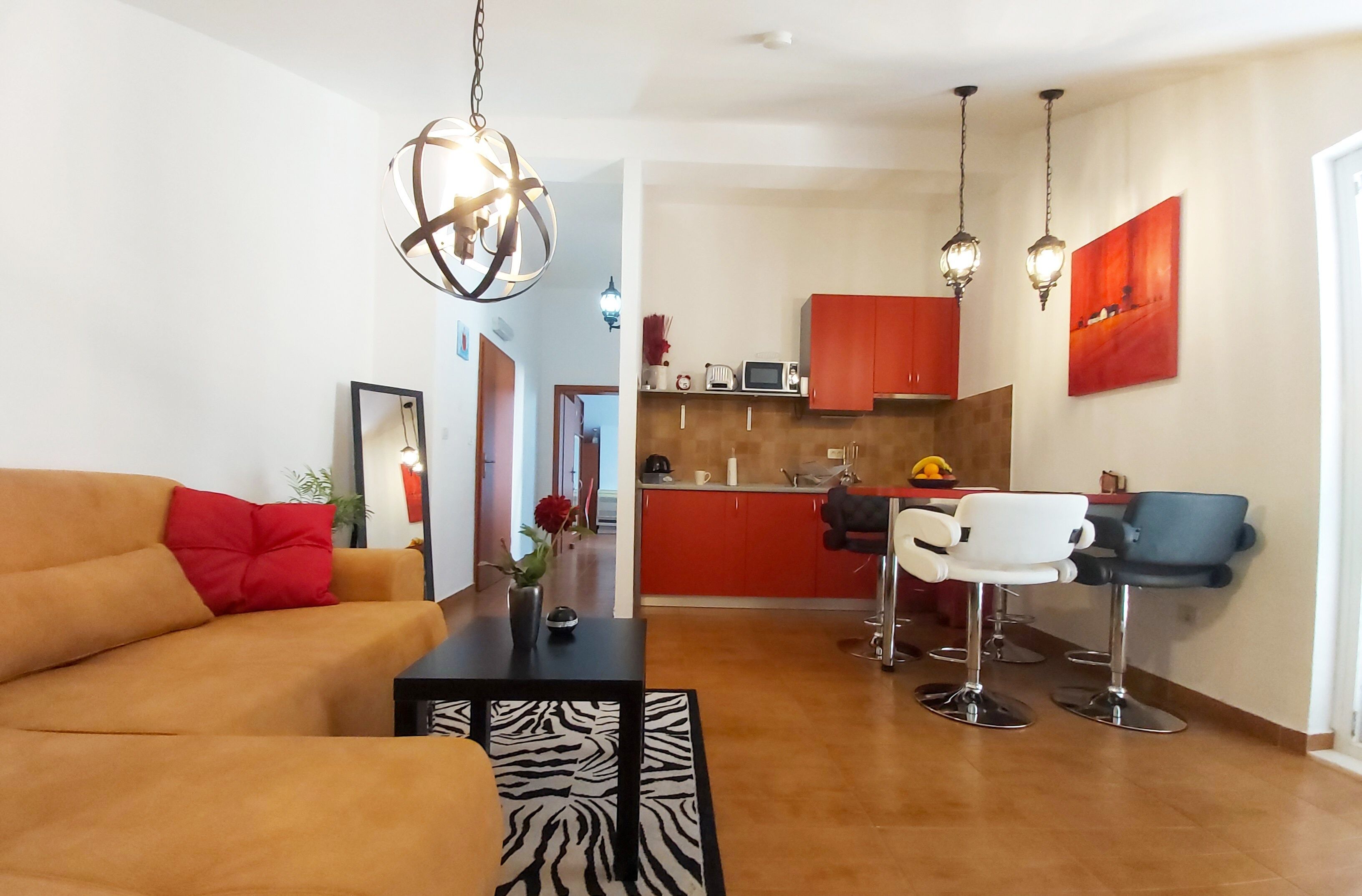 A modern living room with a sofa, a coffee table, and a zebra-patterned rug. There is a kitchen area with red cabinets and a dining area with bar stools.