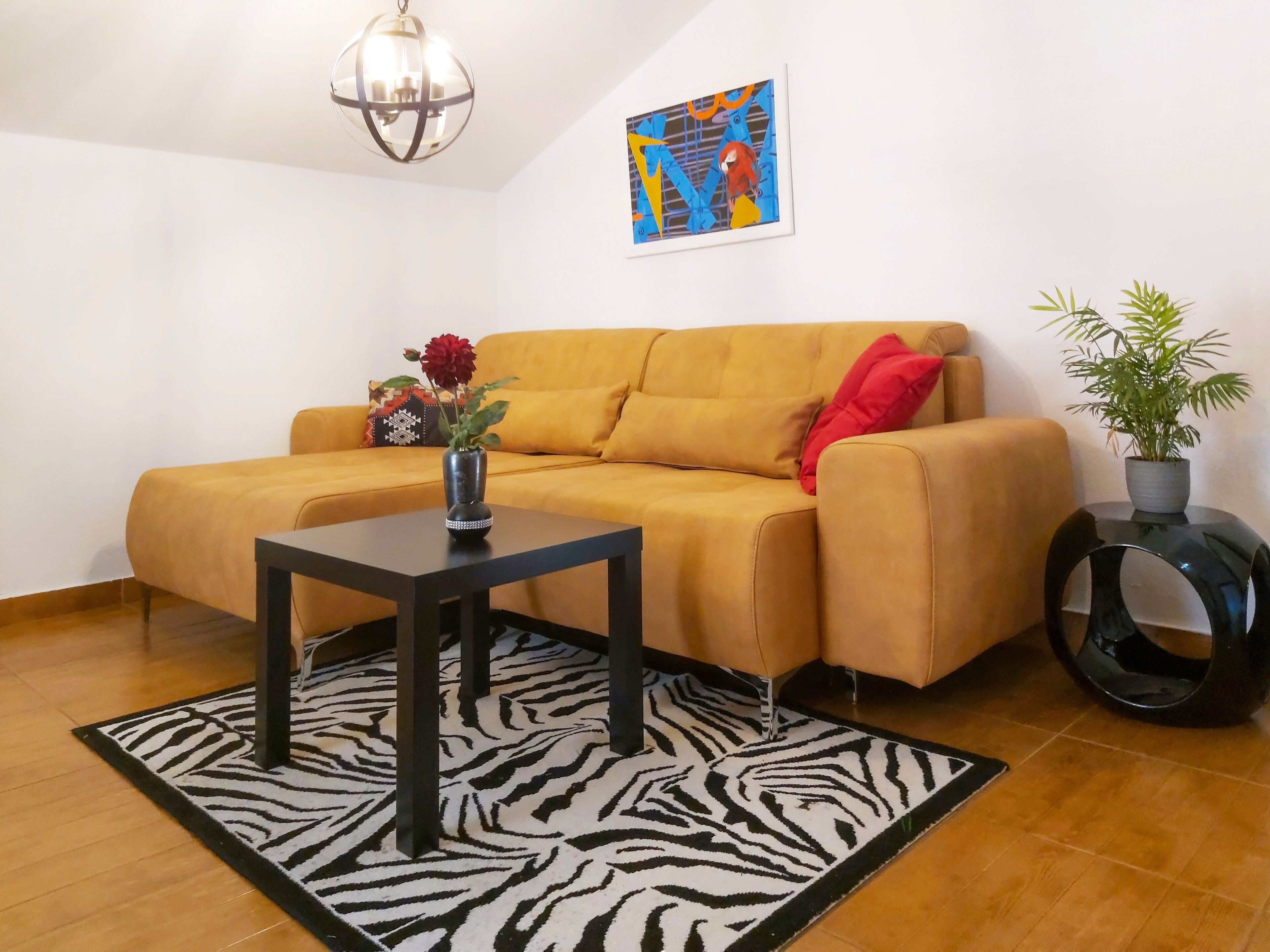 A living room with a yellow sofa, a black coffee table, a zebra-patterned rug, a potted plant, and a wall art piece.