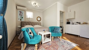 Un dormitorio compacto con cama, dos sillones a juego, una mesita, un espejo decorativo y una kitchenette.