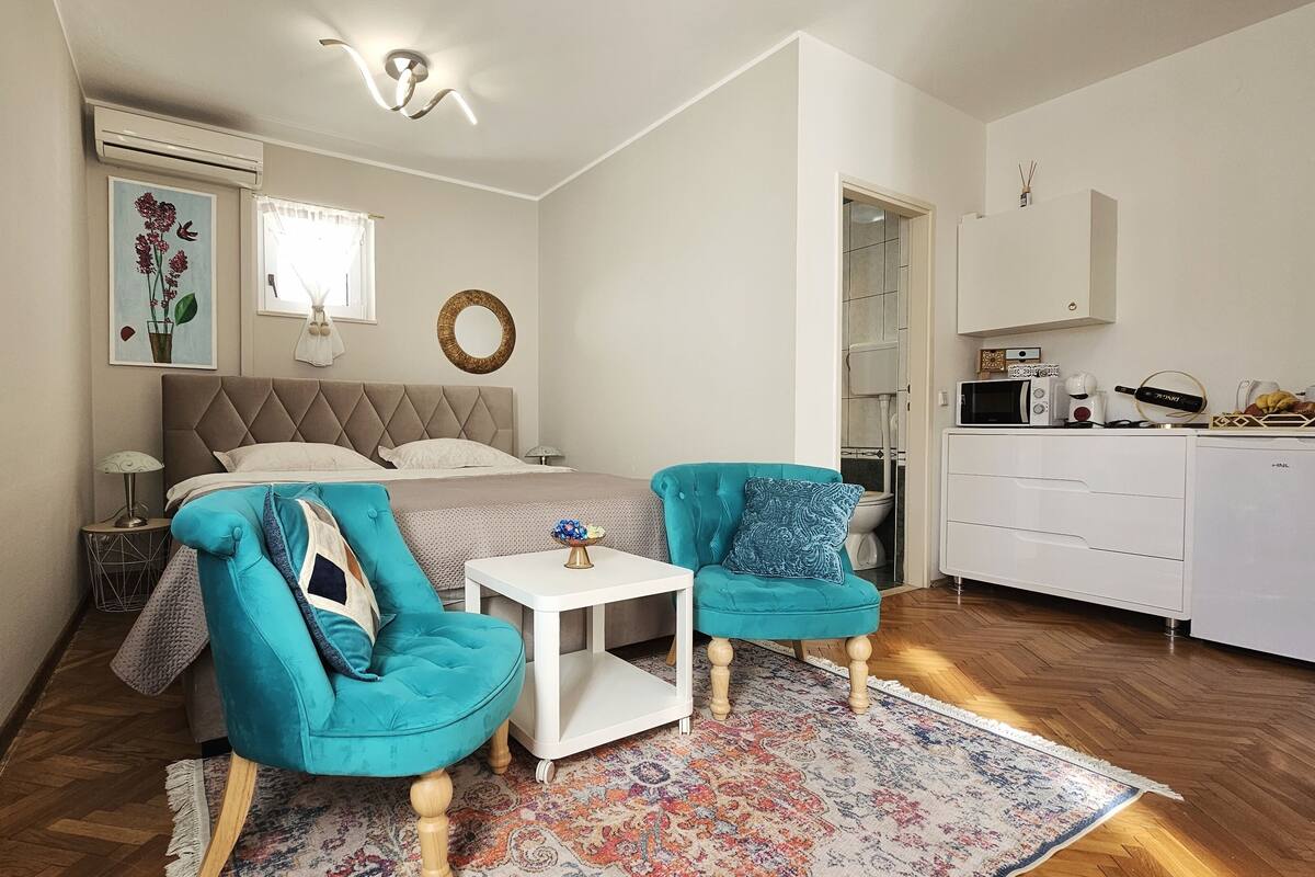 A compact bedroom with a bed, two matching armchairs, a small table, a decorative mirror, and a kitchenette.