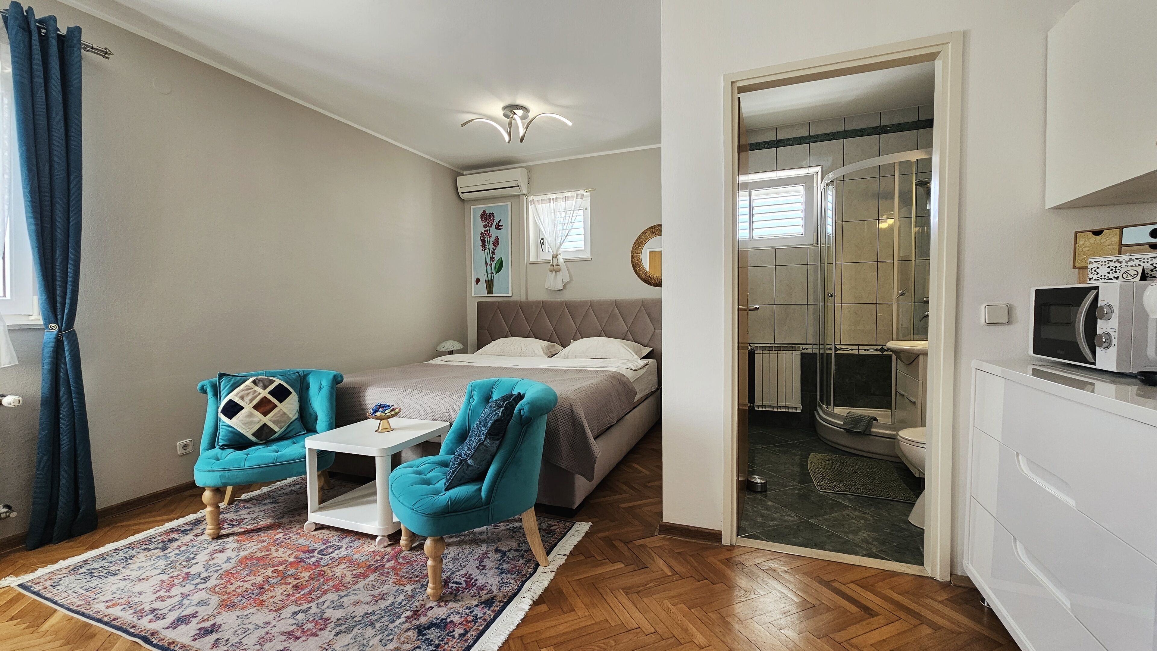 A compact bedroom with a bed, two chairs, a small table, and a shower area visible through an open door.