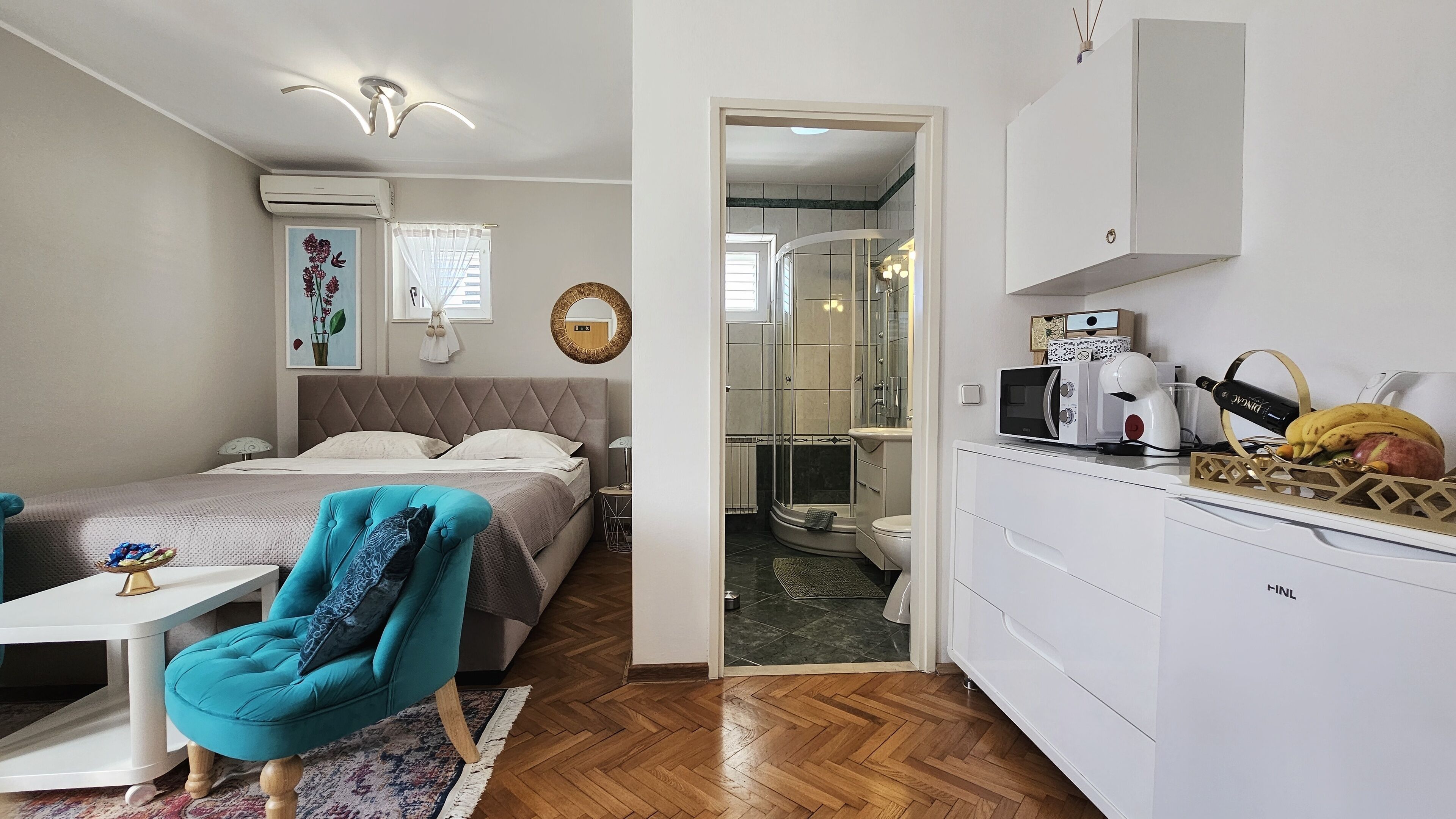 A compact bedroom with a bed, a blue armchair, and a bedside table. A bathroom with a shower and toilet is visible through an open door.