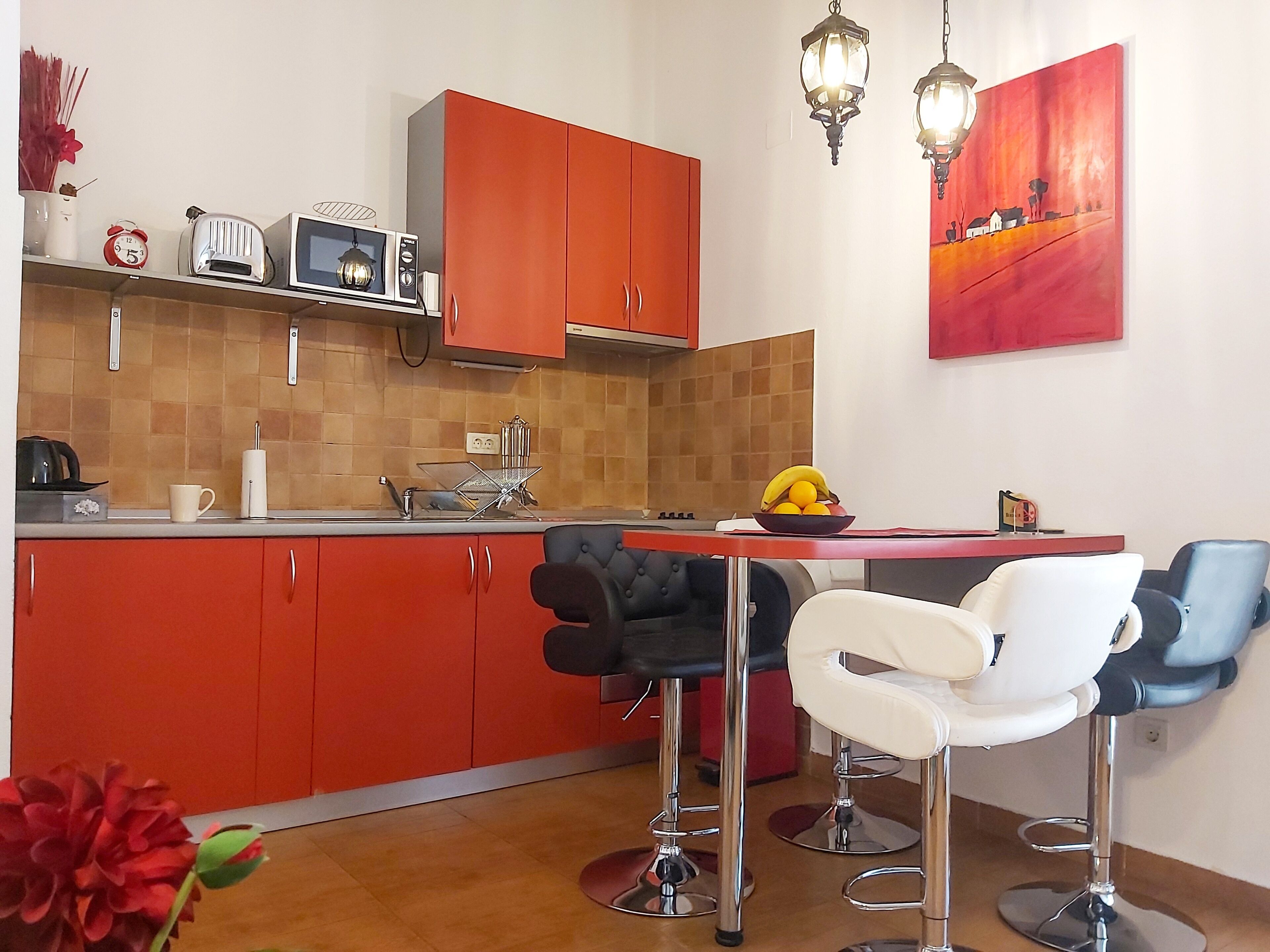 Una cocina compacta con armarios rojos, un microondas, un tostador y un tazón de fruta sobre la encimera. Hay dos taburetes de bar y una mesa con sillas.