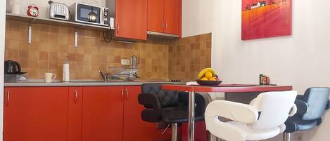 Una cocina compacta con armarios rojos, un microondas, un tostador y un tazón de fruta sobre la encimera. Hay dos taburetes de bar y una mesa con sillas.