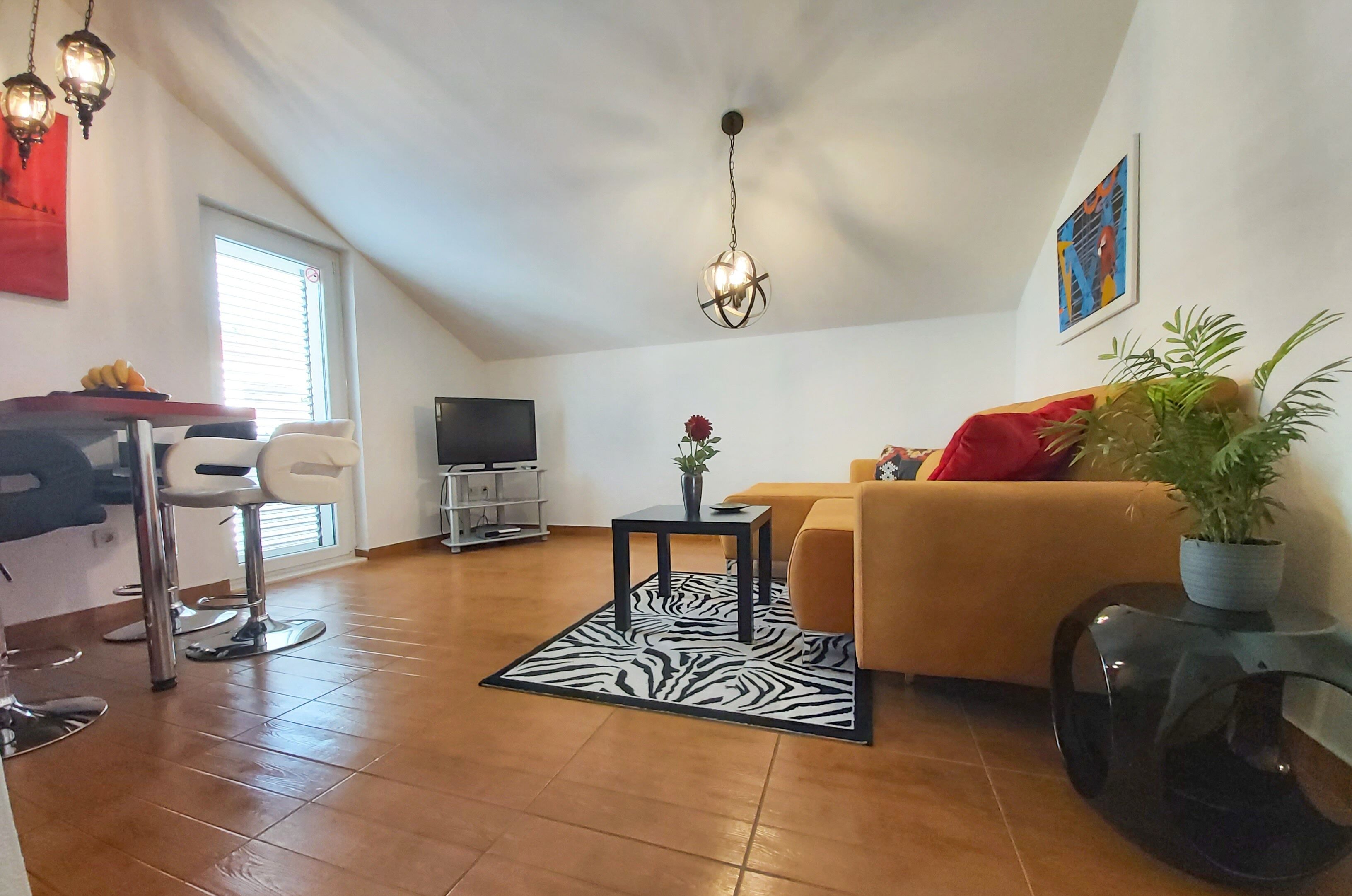 A living room with a sofa, a dining area with chairs, a TV, and a ceiling light fixture.