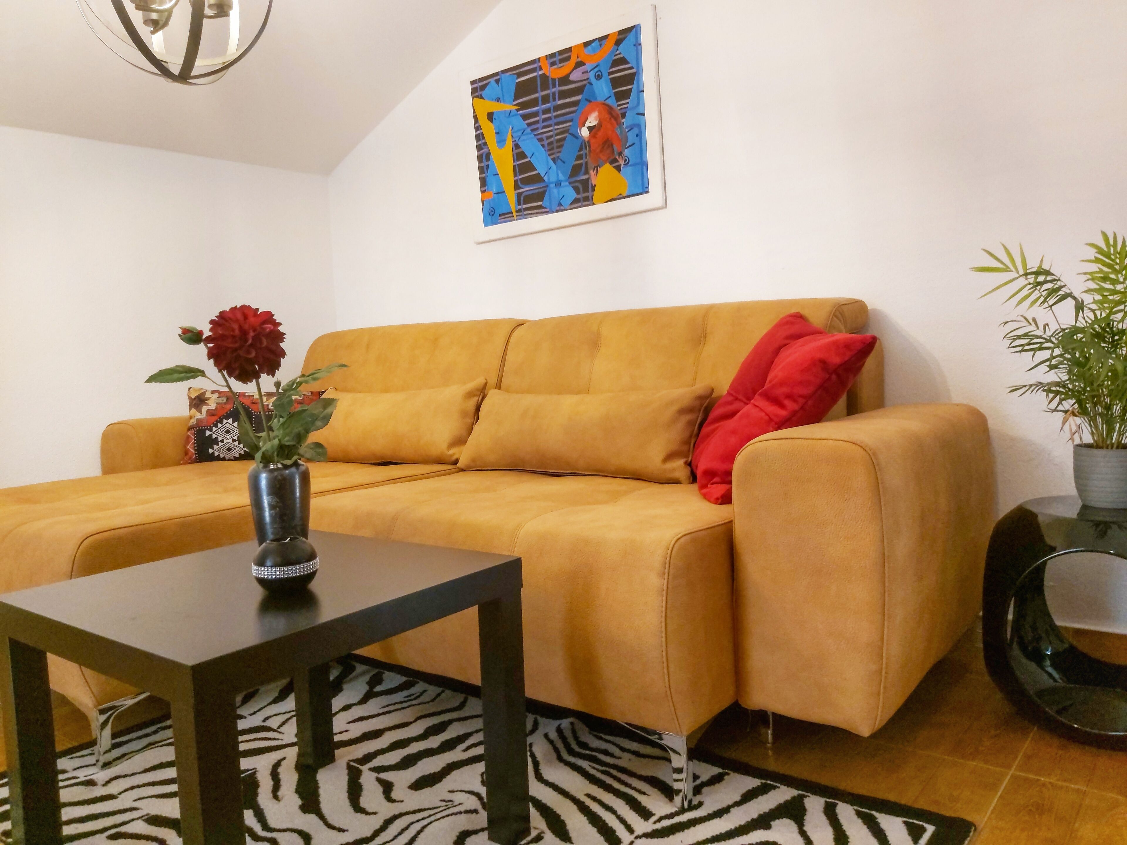 Una sala de estar con un sofá amarillo, una mesita, un florero con una flor roja, un tapete con estampado de cebra y un cuadro en la pared.