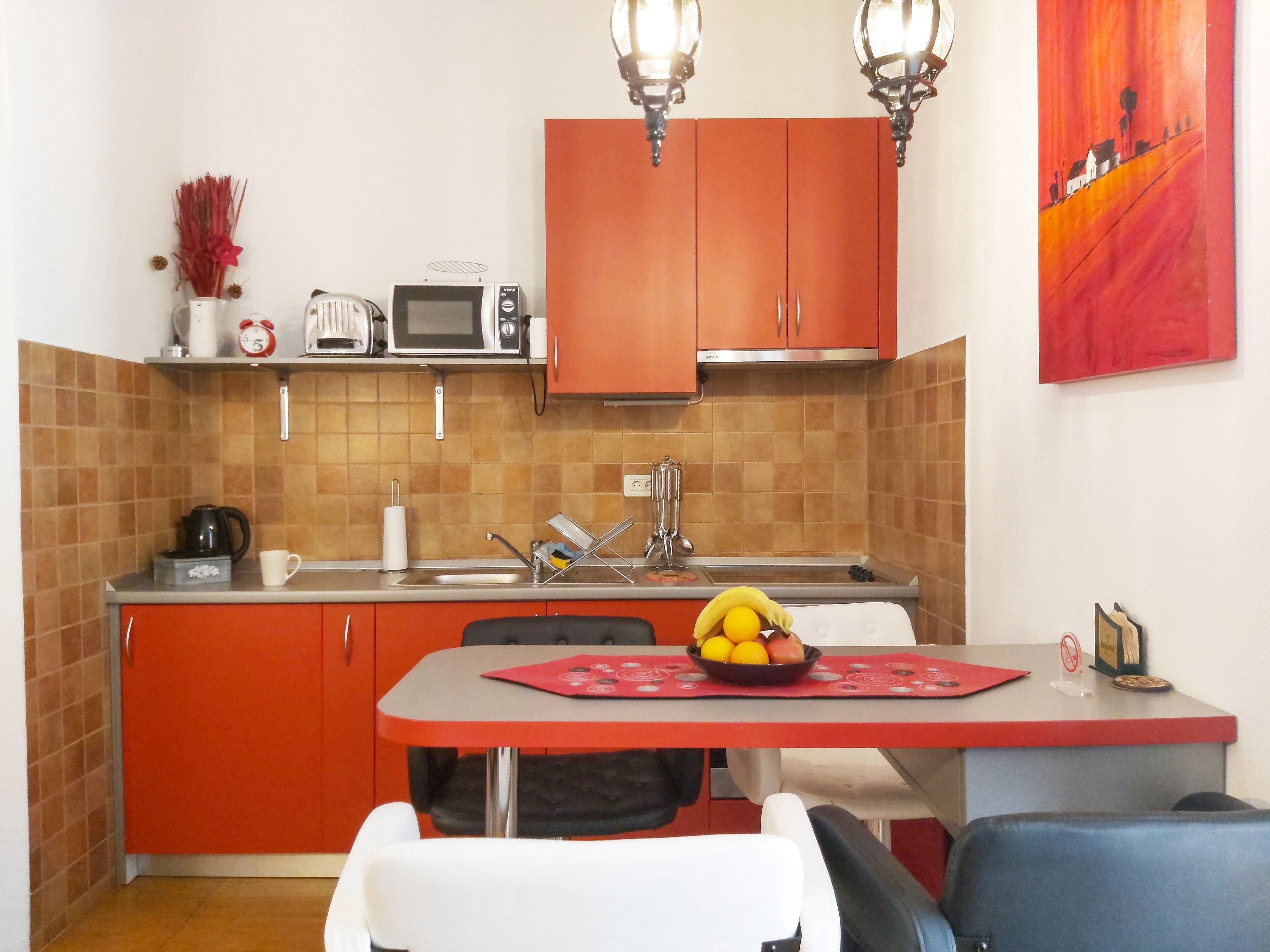 Een kleine keuken met oranje kastjes, een magnetron en een spoelbak. Er staat een eettafel met stoelen en een schaal fruit erop.
