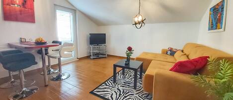 Ein gemütliches Wohnzimmer mit einem gelben Sofa, einem Teppich mit ZEBRA-Muster und einem Fernseher auf einem Schrank. Zwei Sessel, ein kleiner Tisch mit einem Blumenstrauß und ein Bild an der Wand ergänzen das Zimmer.