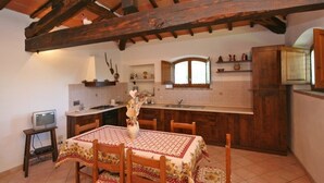 A rustic kitchen with wooden beams, a tiled floor, a dining table with a floral tablecloth, and a small TV on a stand. - Pienza B - Apartment with pool (Monticchiello)