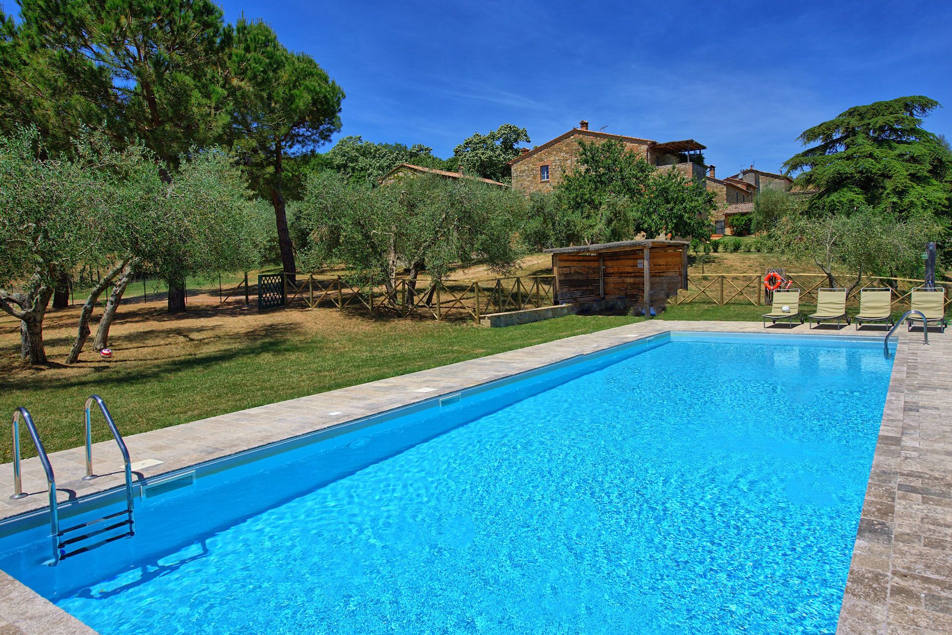 A rectangular swimming pool with a clear blue water surface, surrounded by a stone deck. There are metal pool ladders on the left side. In the background, there are trees, a wooden structure, and a stone building with a red roof.
