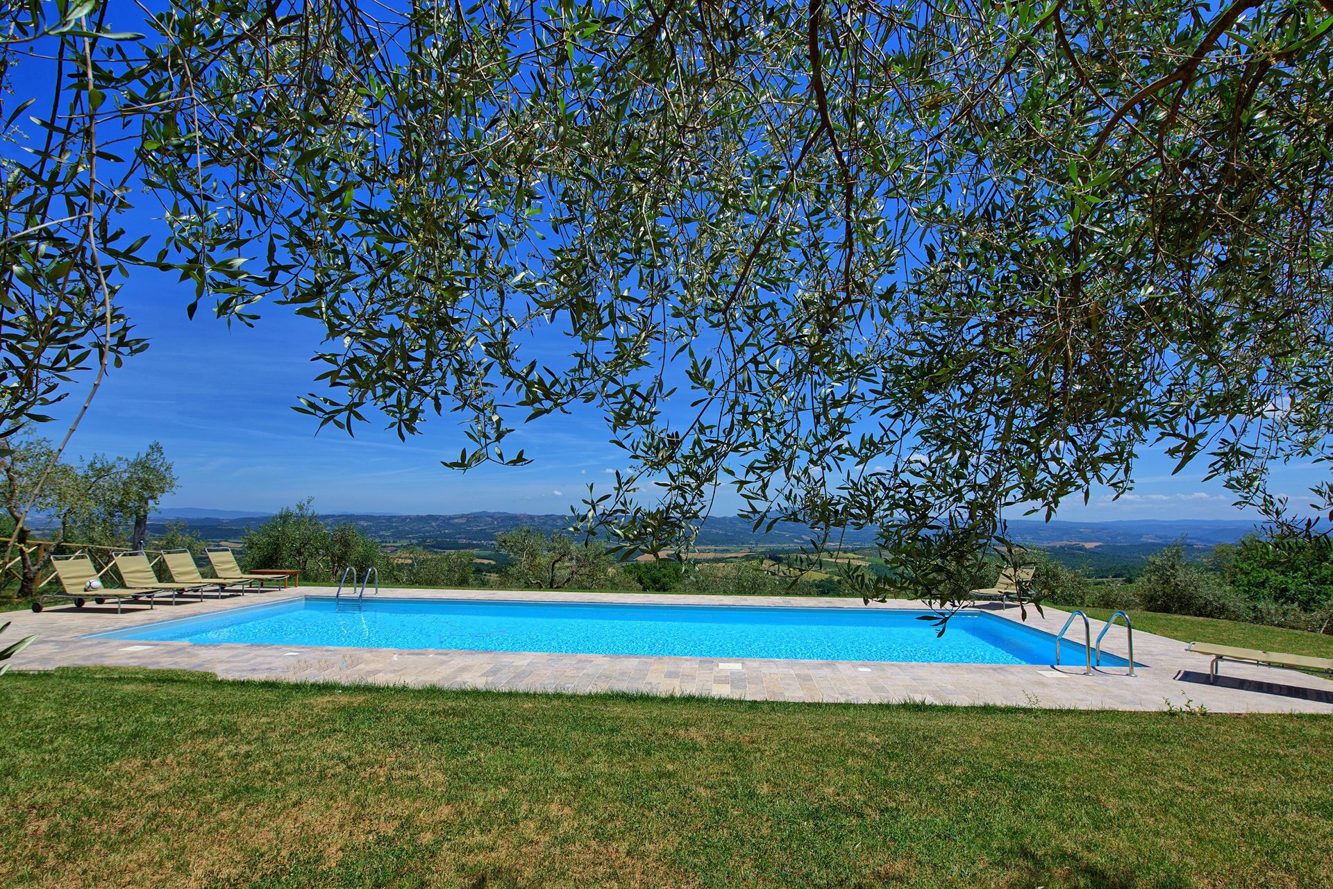 A rectangular swimming pool with a clear blue water surface, surrounded by a stone deck. There are lounge chairs and a small table on the deck. The pool is set against a backdrop of a clear blue sky and a distant landscape.