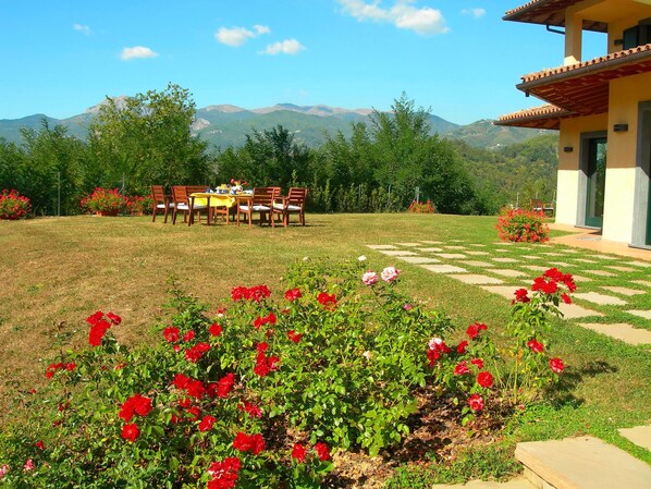 A dining area with a table and chairs, surrounded by a garden with red flowers and a view of mountains.