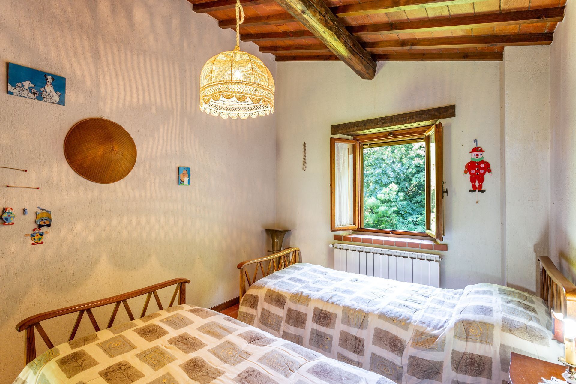 Une chambre avec deux lits en bois, une lampe suspendue, une fenêtre donnant sur un paysage verdoyant et un radiateur.