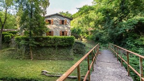 Ein Steinhaus mit Holzhörnern, ein Kiesweg führt hinauf, und eine Holzbrücke überspannt einen Bach.