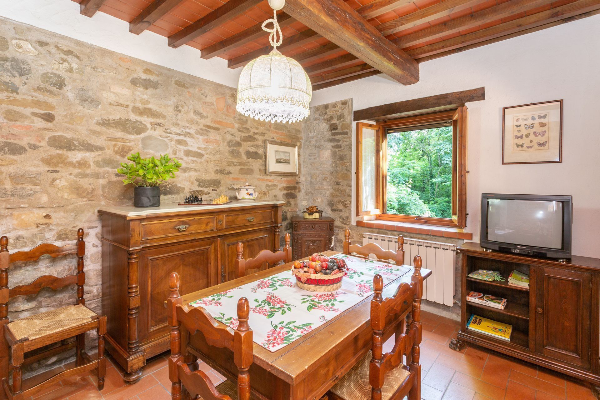 Un comedor rústico con una mesa de madera, sillas, una araña, una pared de piedra, un ventanal con vistas a la vegetación y un televisor sobre un mueble de madera.