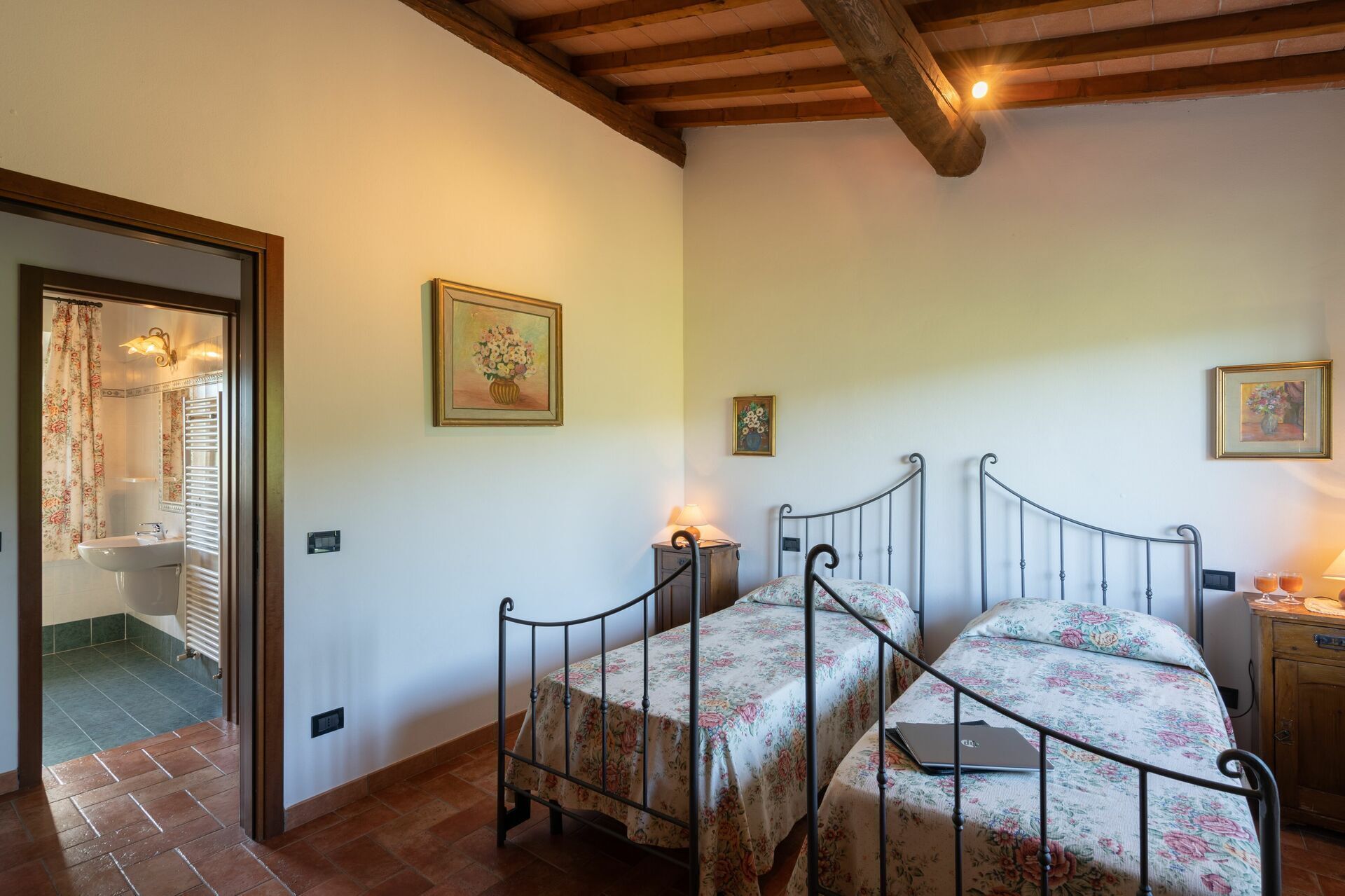 A bedroom with two single beds, a wooden headboard, a small bedside table with a lamp, and a bathroom visible through an open door.