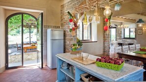 A kitchen with a stone wall, a refrigerator, a dining area with white chairs, and a fruit basket on the counter. - Villa Sant' Angelo - Villa with private pool (Poppi)