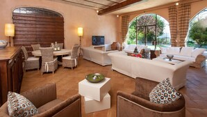 A spacious living room with a large sofa, two armchairs, a dining table with wicker chairs, a TV, and a large arched window offering a view of greenery. - Villa Gubbio - Luxury Villa Rental with private swimming pool in Gubbio, Umbria (Gubbio)