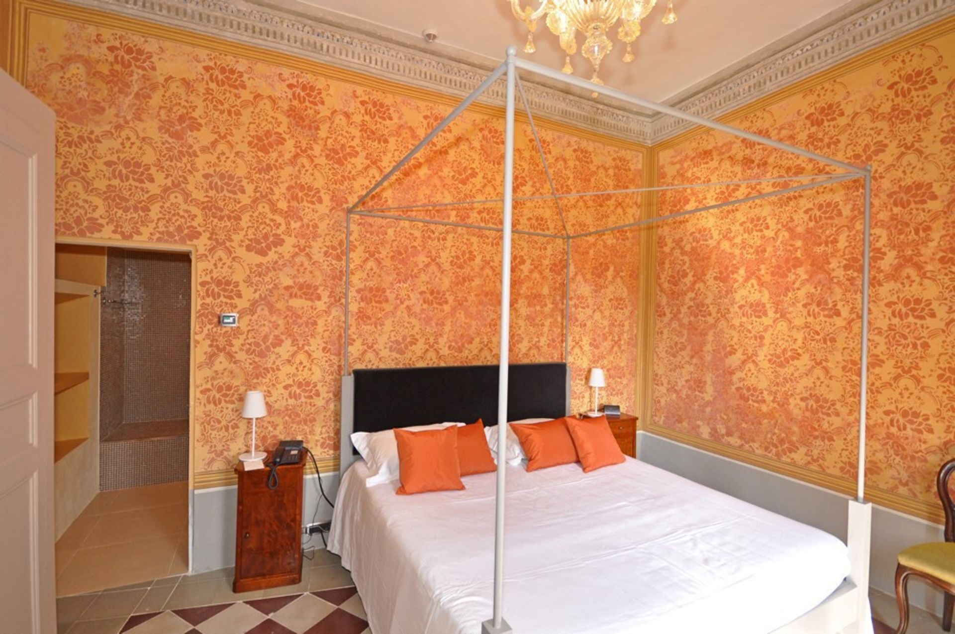 A four-poster canopy bed with white bedding and orange pillows, a wooden nightstand with a lamp, and a telephone.
