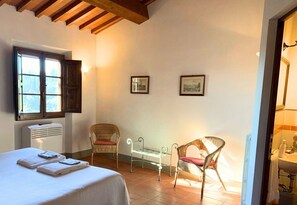Une chambre d’hôtel avec un lit, deux fauteuils en osier, une petite table, une fenêtre donnant sur un paysage agréable et deux tableaux encadrés accrochés au mur.