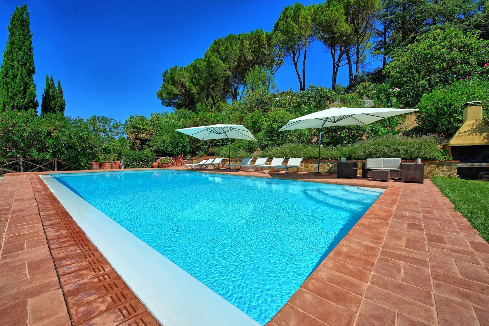 A rectangular swimming pool with clear blue water, surrounded by a brick patio. There are white lounge chairs and umbrellas along the poolside.
