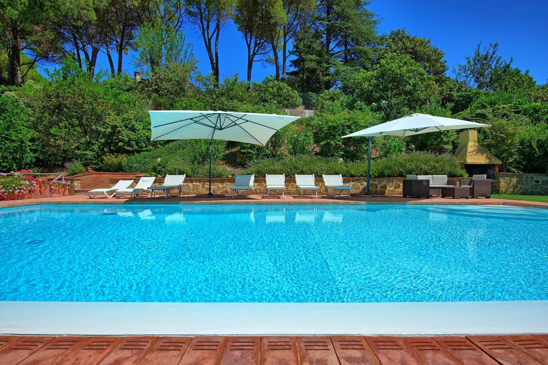 A swimming pool with clear blue water, surrounded by lounge chairs and umbrellas, set against a backdrop of trees and a stone wall.