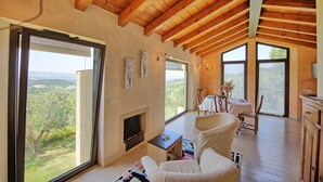 Amplia sala de estar con chimenea, ventanas grandes que ofrecen vistas al campo y vigas de madera en el techo.