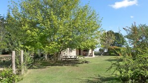 Un edificio in pietra con un tavolo da picnic davanti, immerso nel verde di alberi rigogliosi e sotto un cielo azzurro limpido.