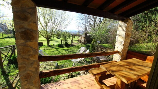 Un terrazzo in legno con tavolo e sedie da picnic, affacciato su una zona erbosa con alberi e una vista lontana delle colline.