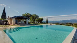 Una piscina con agua cristalina azul, un área de descanso con sillas, una pequeña construcción con techo de paja y vistas a montañas distantes.