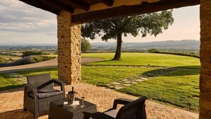Un patio al aire libre con mobiliario de mimbre y un pilar de piedra, con vistas a un paisaje cubierto de césped y colinas distantes.