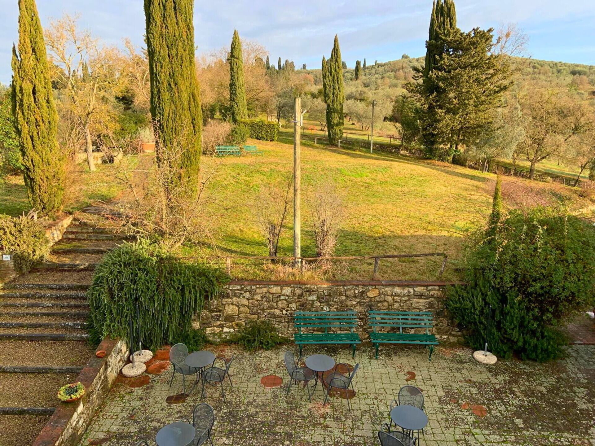 Une cour avec des marches en pierre menant à un espace aménagé avec des tables et des chaises en métal, le tout entouré de verdure et d’arbres.
