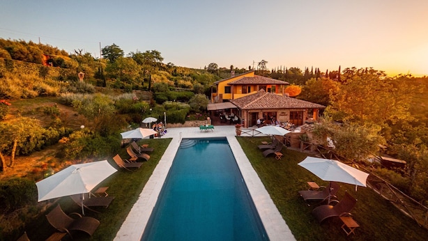 Une piscine rectangulaire, agrémentée de chaises longues et de parasols, entourée d’un jardin aménagé et d’un bâtiment dont le toit est recouvert de tuiles.