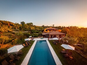 Une piscine rectangulaire, agrémentée de chaises longues et de parasols, entourée d’un jardin aménagé et d’un bâtiment dont le toit est recouvert de tuiles.