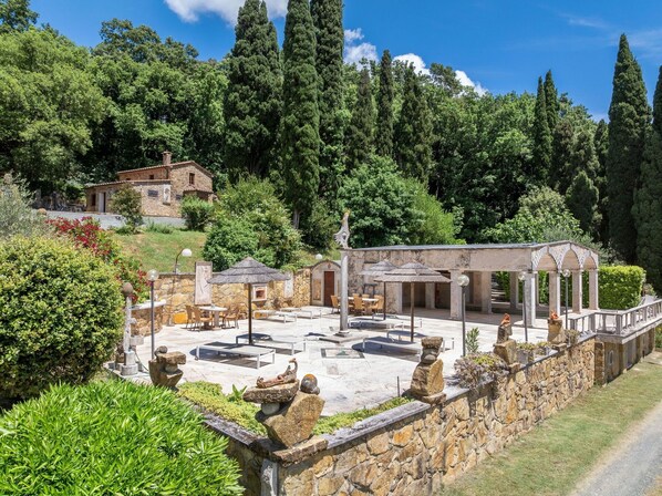 A stone building with outdoor seating and a pergola, surrounded by greenery and trees.