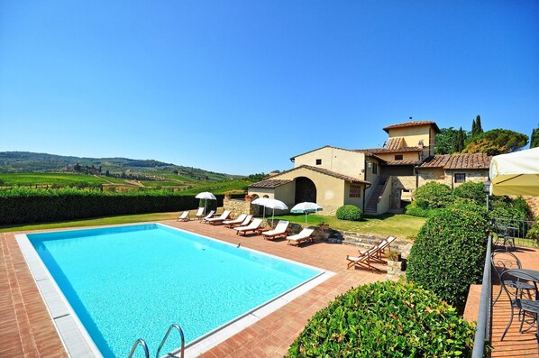 A swimming pool with lounge chairs and umbrellas, surrounded by a landscaped garden and a building with a tiled roof.