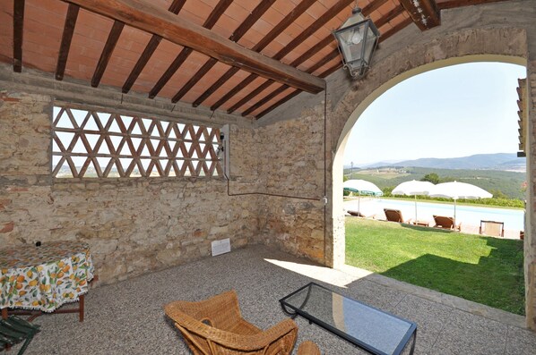 A stone-walled room with a wooden ceiling, a lattice window, a table with a fruit pattern tablecloth, a wicker chair, and a glass table. Outside, there are umbrellas and a pool with a view of hills.