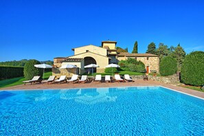A poolside area with lounge chairs and umbrellas, a stone building with a tiled roof, and a clear blue sky.