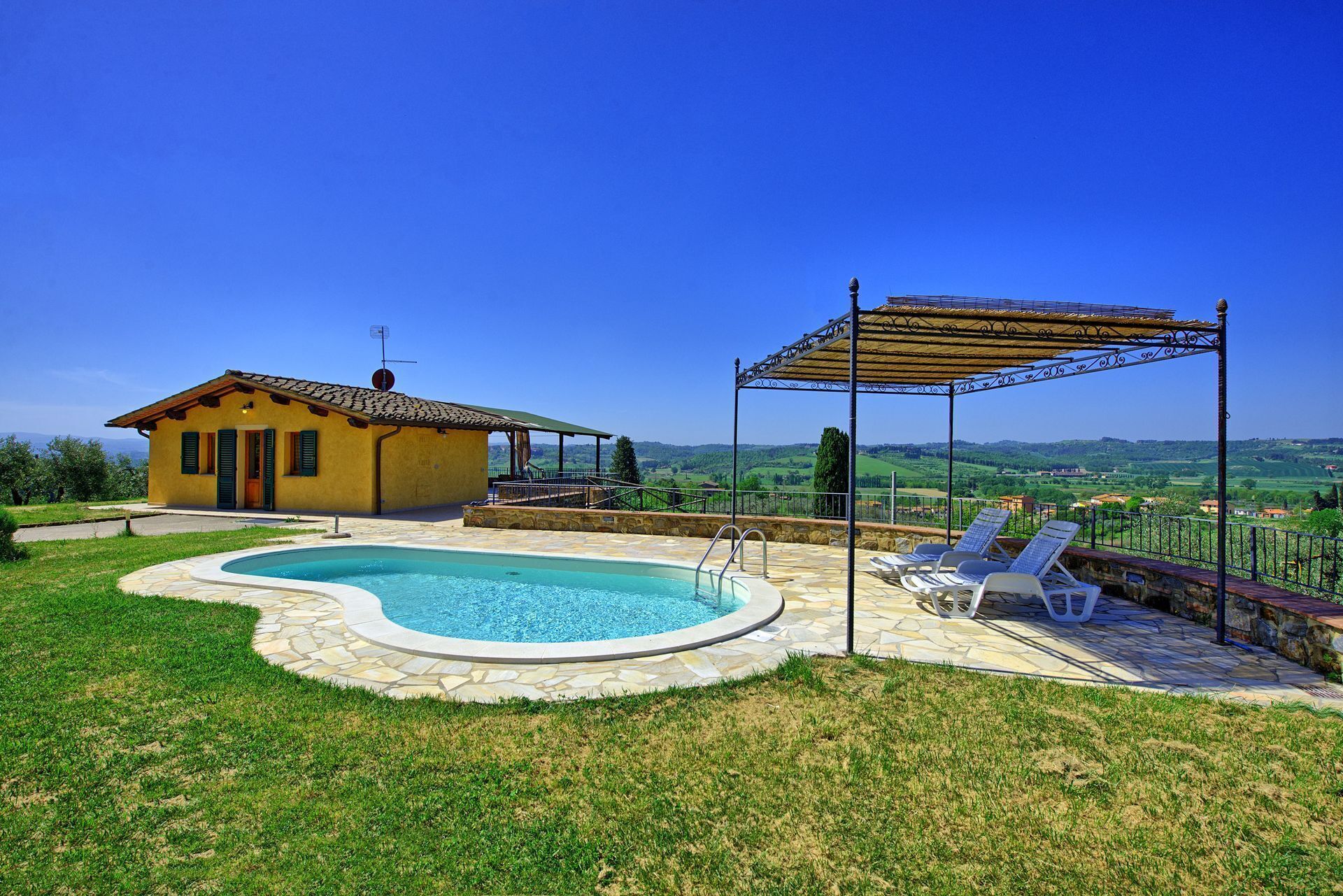 Ein Poolbereich mit Steinplatten, einem gelben Gebäude und einer Pergola mit Liegestühlen.