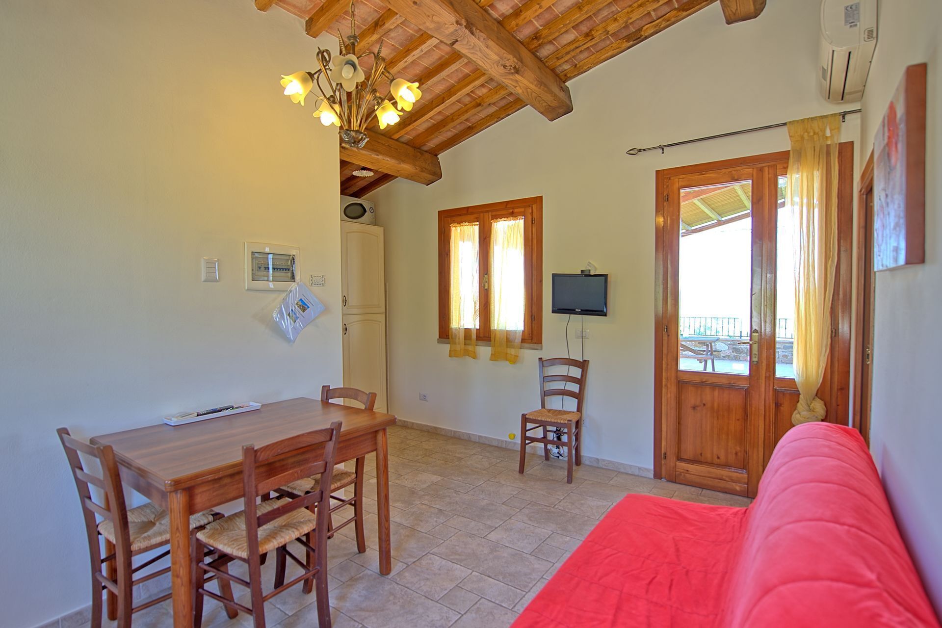A dining area with a wooden table and chairs, a red sofa, a television mounted on the wall, and a wooden ceiling.