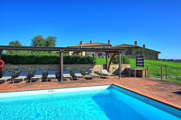 A pool area with lounge chairs, a pergola, and a stone building in the background.