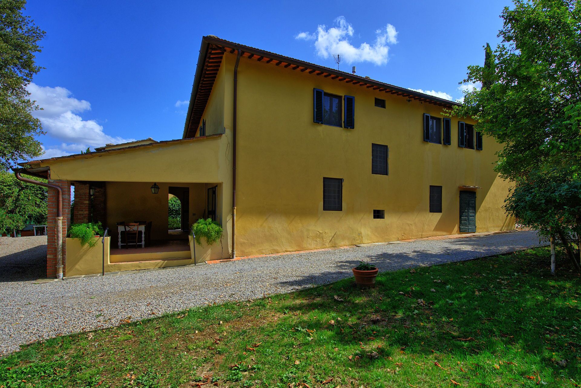 A yellow building with a covered porch, a gravel driveway, and a grassy area with a potted plant.