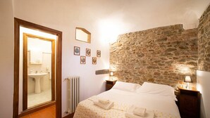 Een slaapkamer met een stenen muur, een bed met witte lakens, een nachtkastje met een lamp, en een badkamer zichtbaar door een open deur.