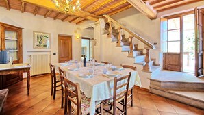 Ruokailutila, jossa on pitkä ruokapöytä katettuna, puiset tuolit, kattokruunu ja portaikko, joka johtaa ylemmälle tasolle.
