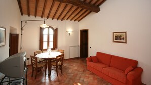 Un comedor con una mesa redonda, sillas, un televisor sobre un mueble bajo, un sofá rojo, una ventana con luz natural y una puerta.