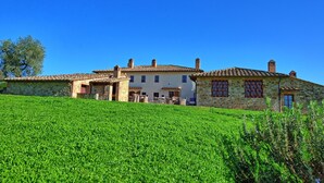 Un edificio de piedra tradicional con techo de tejas, una pequeña estructura adosada con chimenea, y un campo verde exuberante en primer plano.