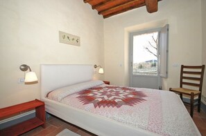 Un dormitorio con una cama, dos lámparas de noche, un banco de madera, una silla y un ventanal con vistas.