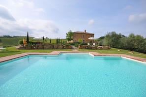 A rectangular swimming pool with clear blue water, surrounded by a stone deck. In the background, there is a stone building with a tiled roof, and a grassy area with trees and shrubs.
