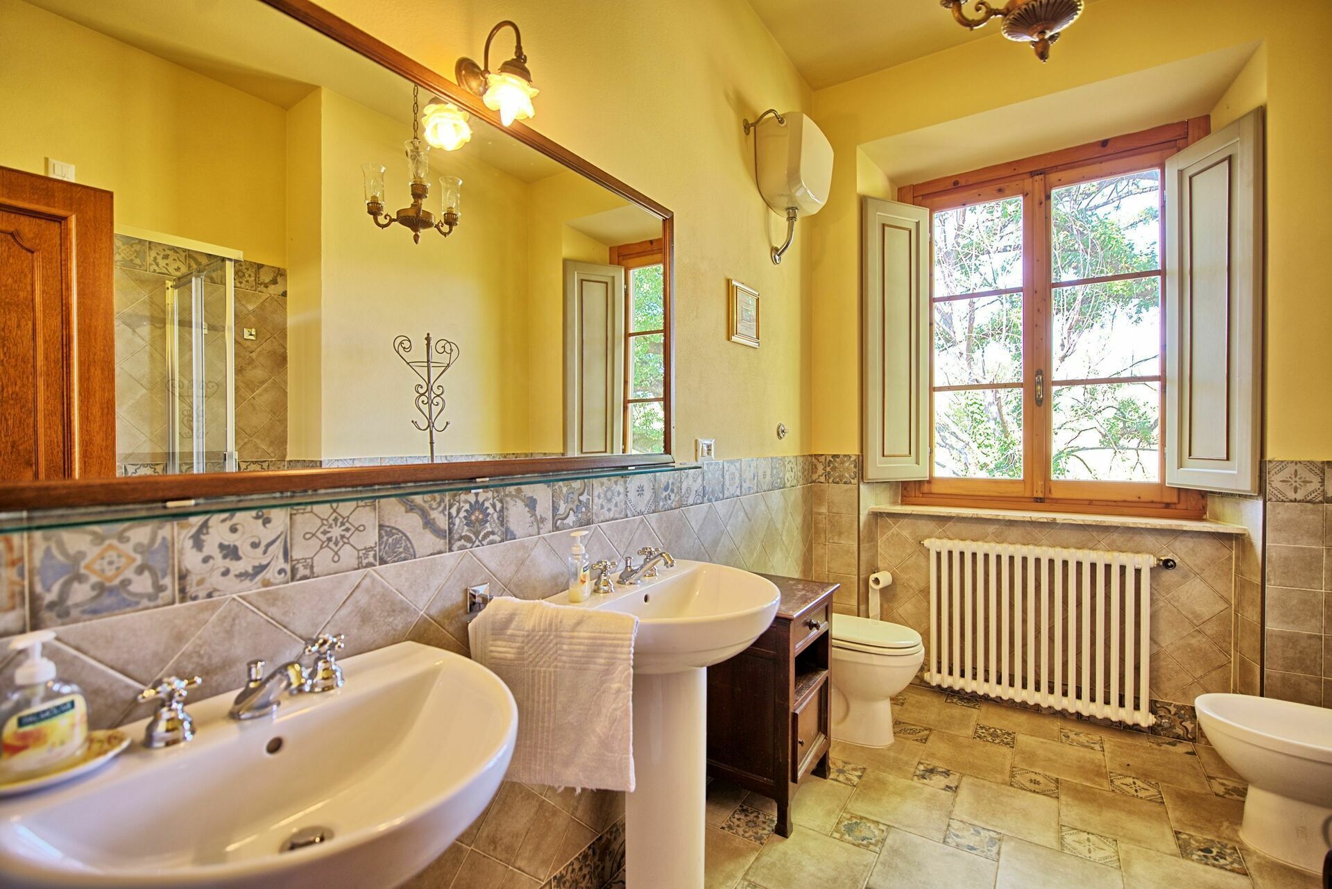 A bathroom with two sinks, a toilet, a radiator, and a window with a view of trees.